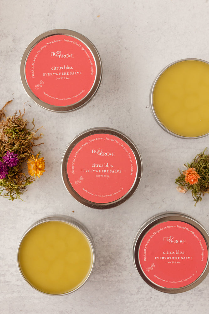 Four tins of Citrus Bliss 'Everywhere Salve' with pink labels on a textured surface with flowers - Fig & Grove
