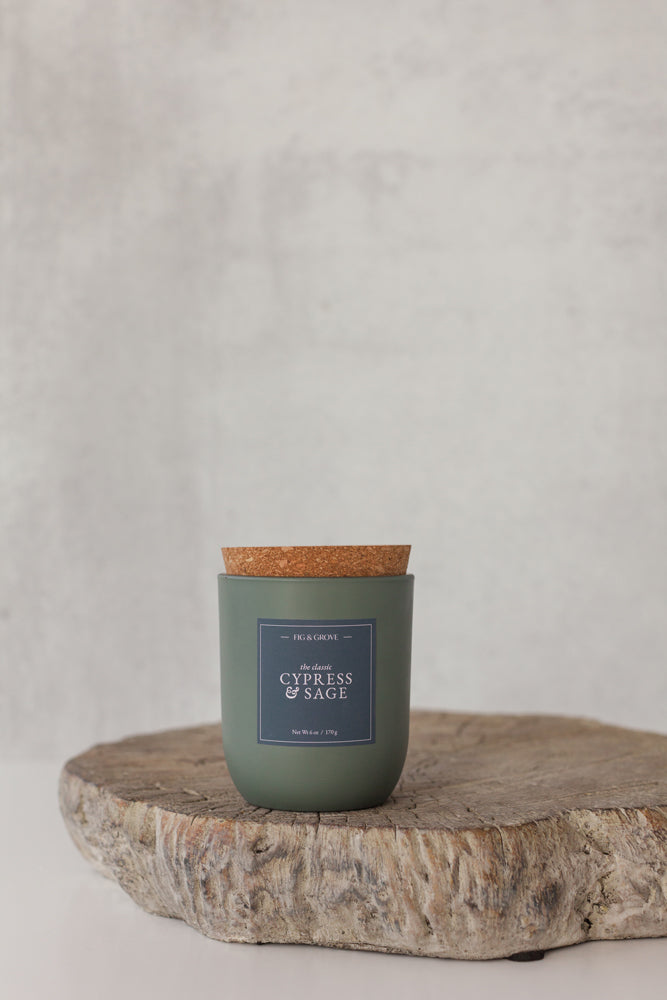 Cypress & Sage Candle in a sage green glass jar with cork lid on a wooden surface against a light gray background