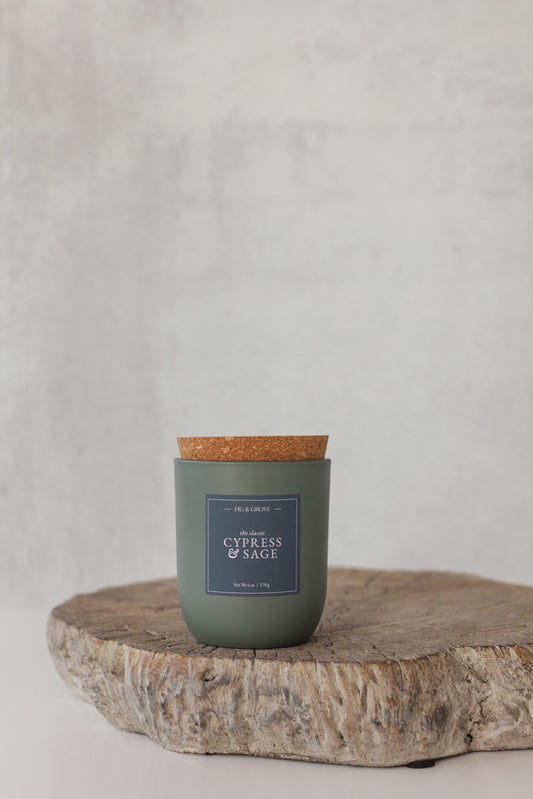 Cypress & Sage Candle in a sage green glass jar with cork lid on a wooden surface against a light gray background